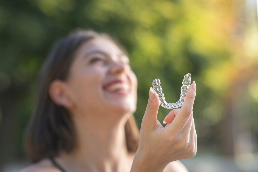 Close-Up of Clear Aligners Held by Smiling Woman – Invisalign in Cherry Creek A woman happily displays her transparent aligners against a bright outdoor background, highlighting the aesthetic and functional benefits of modern clear aligner therapy – Invisalign in Cherry Creek.