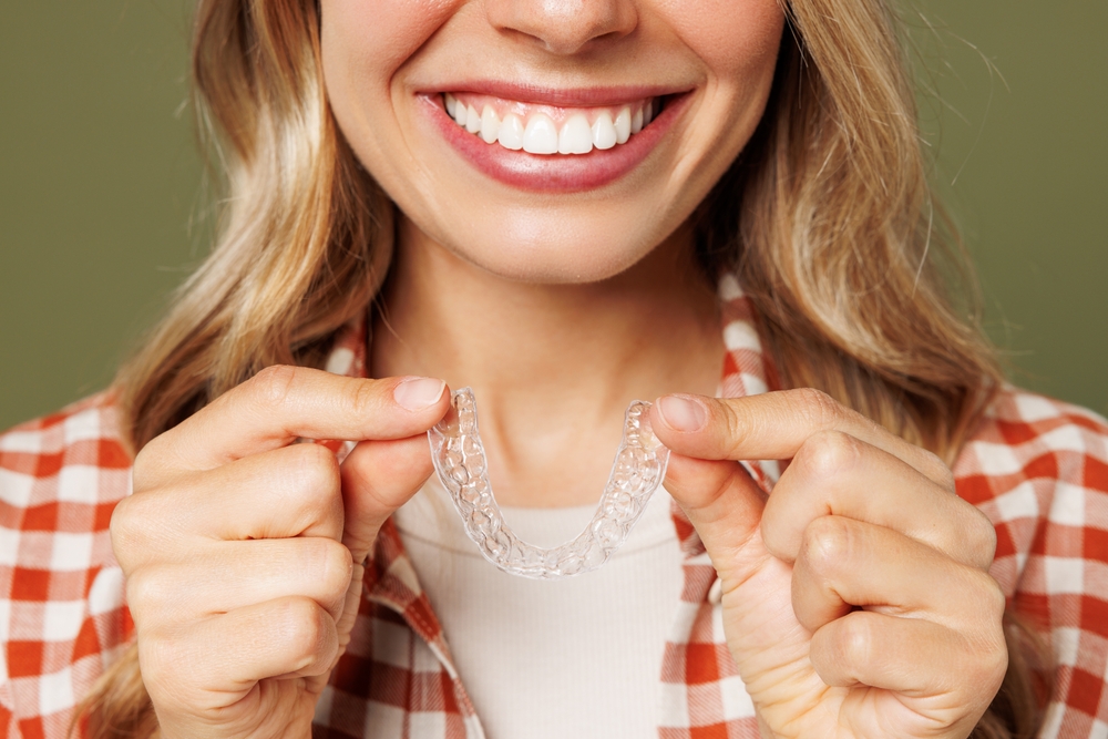 Woman Smiling While Holding Clear Aligner – Invisalign in Highlands A woman shows off a bright, confident smile while holding a clear aligner tray, highlighting the convenience and subtlety of modern orthodontic treatment – Invisalign in Highlands.