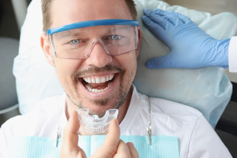 Male Patient Smiling with Custom Night Guard at Dentist - Night Guards A man in a dental chair smiles as he holds a clear custom night guard, with his dentist nearby ensuring proper fit and comfort. - Night Guards