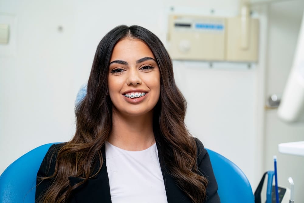Young Woman Smiling in Dental Clinic with Braces - Braces A young woman with long dark hair smiles proudly in a dental office, showing her metal braces while seated in a blue dental chair. - Braces