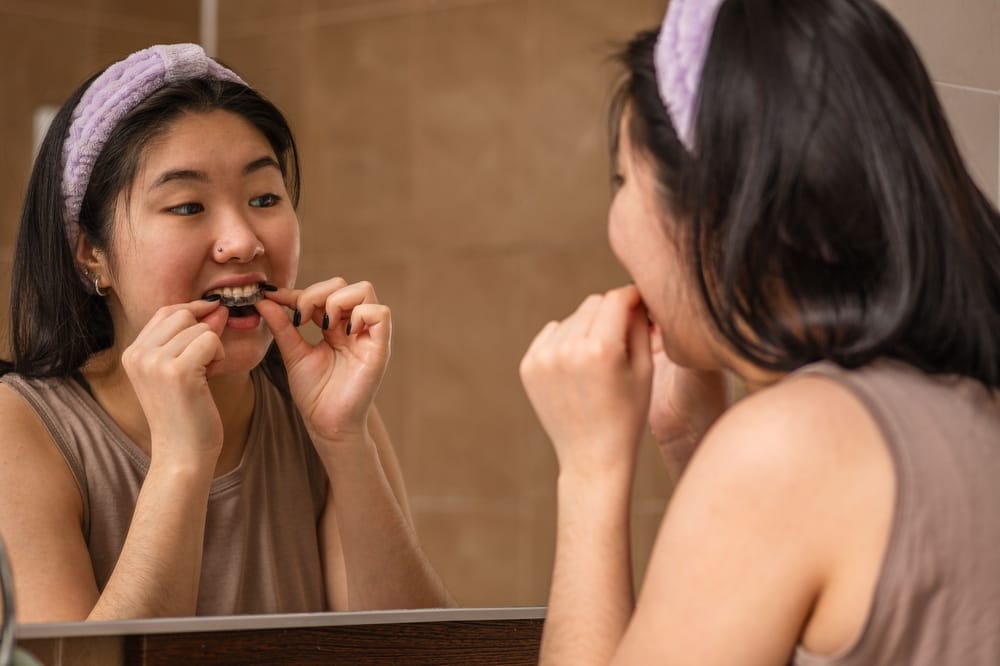 Woman Fitting Night Guard in Front of Mirror - Night Guards A woman stands at her bathroom mirror as she places her night guard on her teeth, demonstrating proper nighttime oral care. - Night Guards