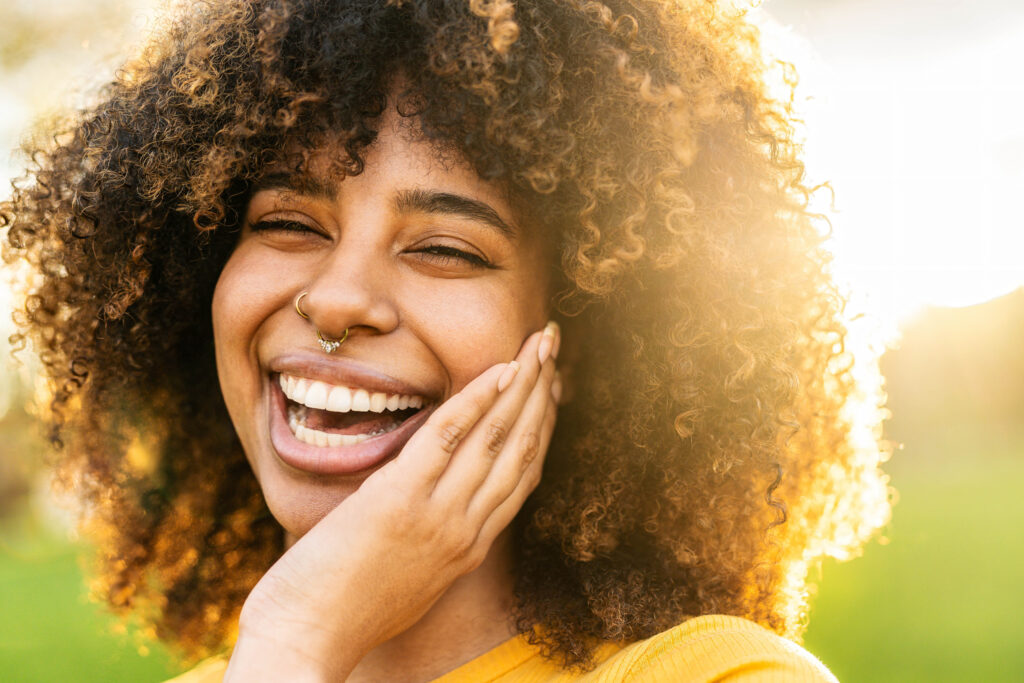 A joyful young woman with curly hair smiles brightly in the warm glow of the sun, showing off her healthy, vibrant smile with confidence – Orthodontics in the Highlands.