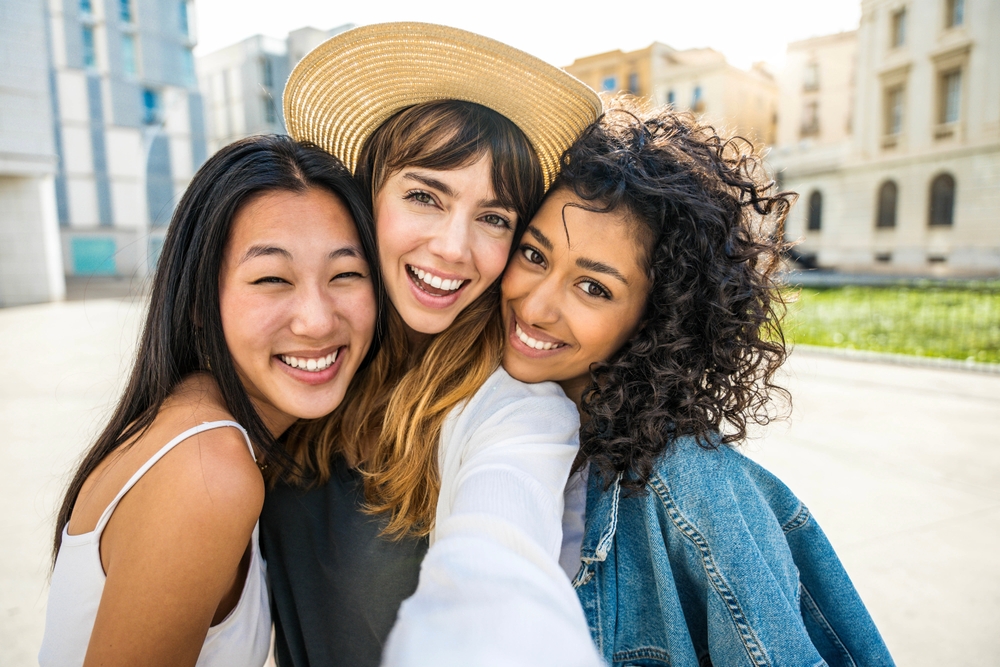 Three friends pose closely for a cheerful outdoor selfie, each showing bright, confident smiles that highlight the joy of shared moments – Orthodontics in the Highlands.