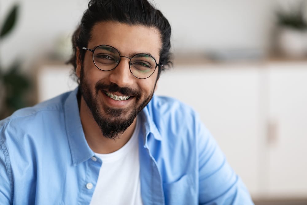 Man with Glasses Smiling with Braces - Braces A cheerful man wearing glasses and a blue shirt smiles warmly, revealing his braces during a relaxed moment at home. - Braces