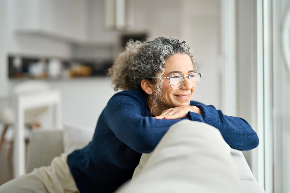 A woman with curly gray hair and glasses smiles softly while leaning on a couch, showcasing comfort and confidence in her healthy, radiant smile – Highland Dental.