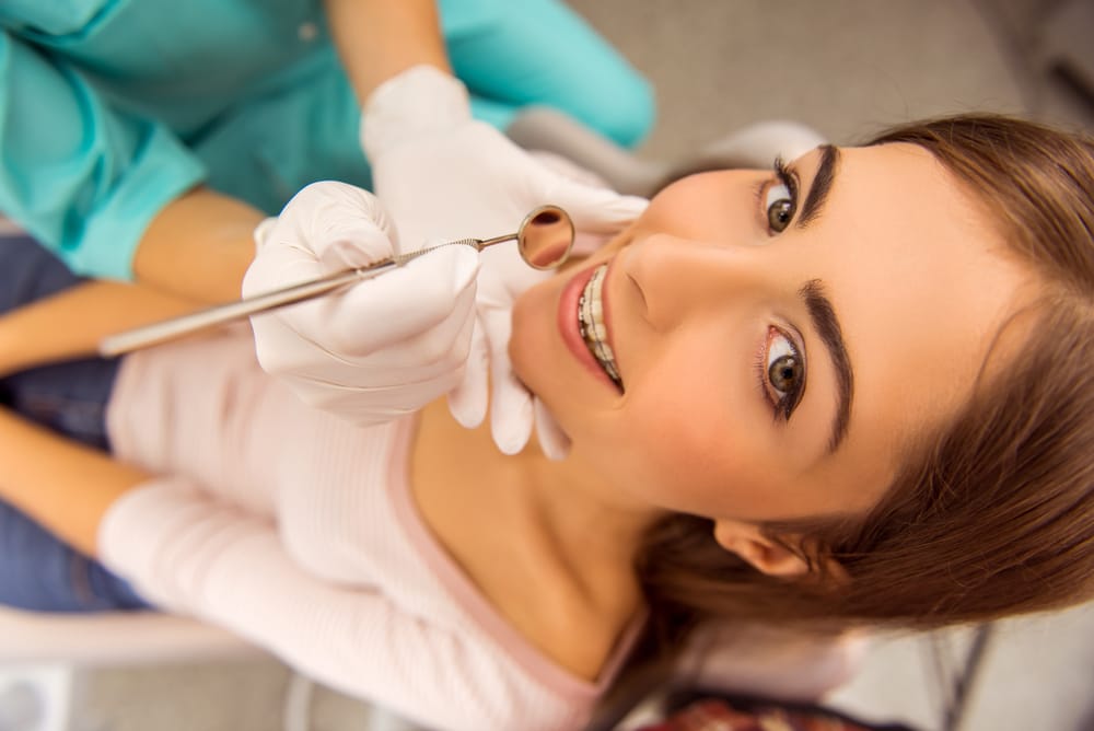 Teen Girl at Dental Checkup with Braces - Braces A teenage girl reclines in a dental chair while a dentist examines her braces with a dental mirror. She smiles confidently during her orthodontic checkup. - Braces