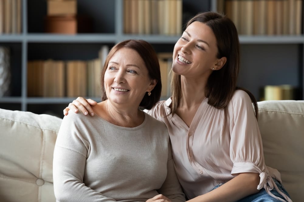 A smiling mother and daughter sit together on a couch, enjoying a warm and joyful moment that highlights their healthy, confident smiles – Orthodontics in Aurora.