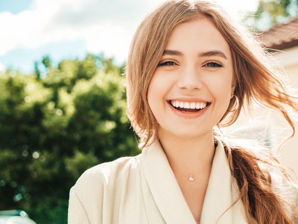 A cheerful young woman smiles brightly while standing outdoors on a sunny day, highlighting her beautifully aligned teeth and vibrant confidence – Orthodontist in Lakewood CO.