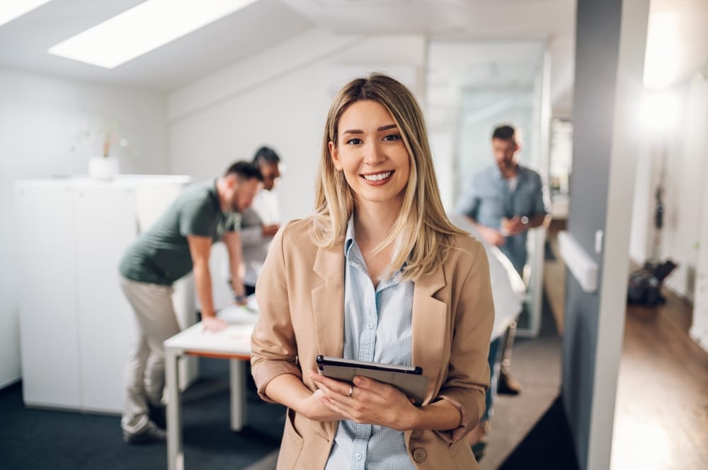 A young professional woman holds a tablet and smiles confidently in a modern office environment, showcasing a bright and well-aligned smile – Orthodontics in Aurora.