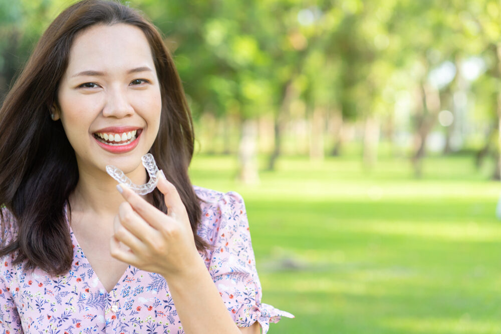 Smiling Woman Holding Clear Aligners Outdoors – Invisalign in Cherry Creek A cheerful woman smiles confidently while holding a set of clear aligners in a sunny park, showcasing the comfort and convenience of discreet orthodontic treatment – Invisalign in Cherry Creek.