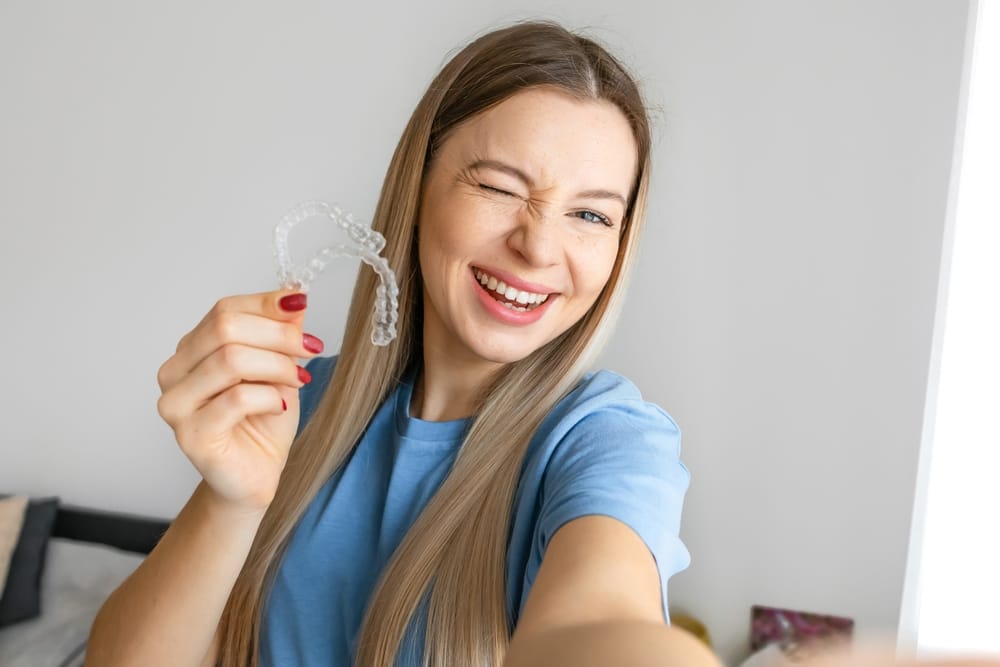 A cheerful woman winks at the camera while holding up a clear retainer, showing off the ease and fun of keeping her smile aligned. - Retainers