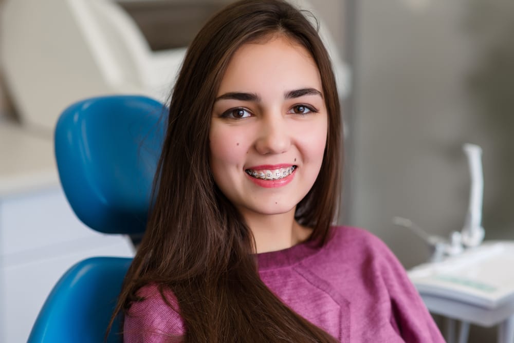 A teen girl with braces smiles while seated in a dental chair, representing professional orthodontic care and steady progress toward a straighter, healthier smile – Braces in Highland.