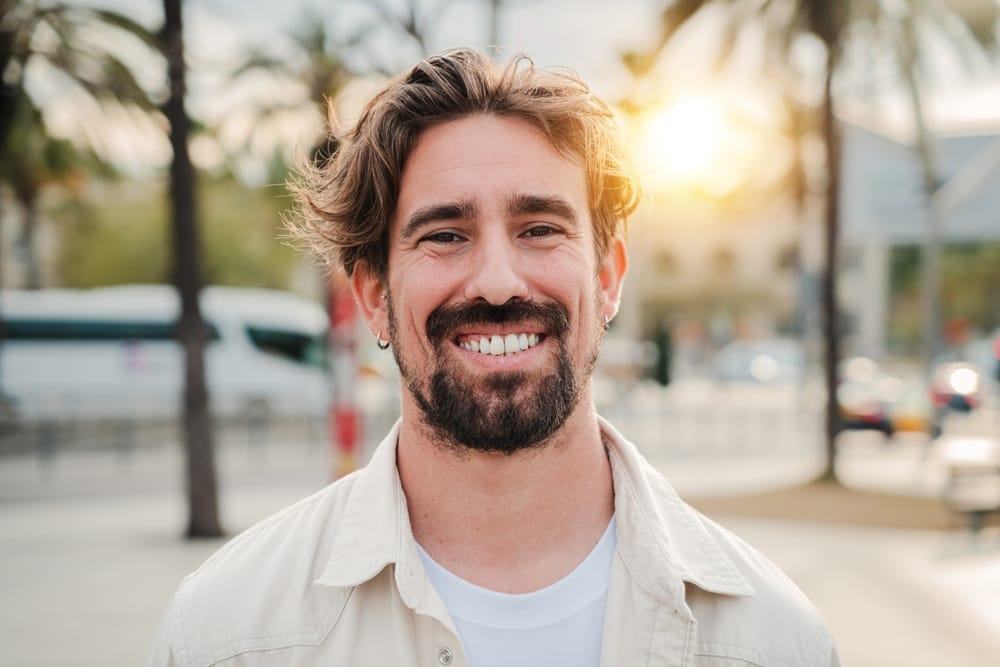 Man Smiling Outdoors After Teeth Whitening – Whitening A cheerful man with a bright, confident smile stands outdoors in natural sunlight, showcasing the results of a professional teeth whitening treatment – Whitening.