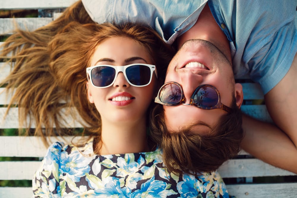 Smiling Couple Relaxing in Sunglasses With Confident, White Smiles – Whitening A happy couple lounges together outdoors wearing sunglasses, each flashing bright, attractive smiles that reflect the benefits of teeth whitening – Whitening.