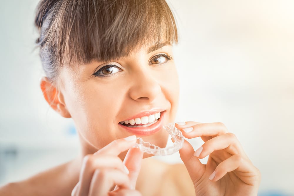 A woman smiles as she prepares to place a clear Invisalign aligner onto her teeth, highlighting the discreet and comfortable nature of removable orthodontics. - Invisalign