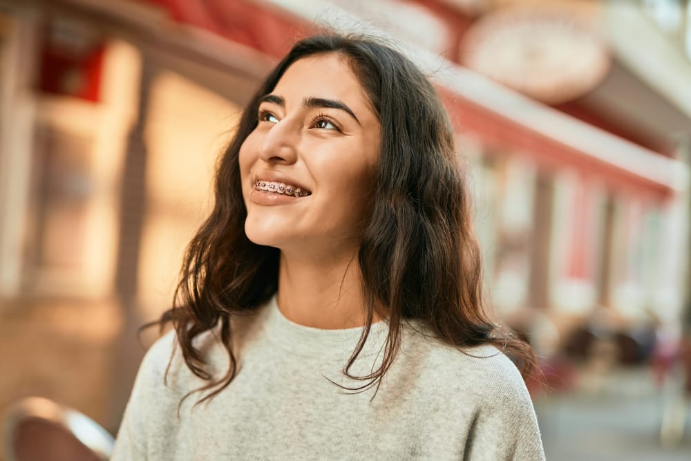 A cheerful young woman smiles confidently while wearing metal braces, showcasing the positive results of orthodontic treatment designed to enhance her smile and bite – Braces in Highland.