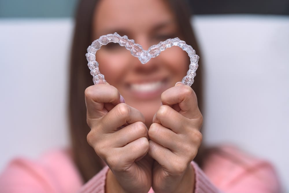 A close-up of a person holding two clear aligners shaped into a heart, symbolizing confidence and love for their smile transformation with Invisalign. - Invisalign