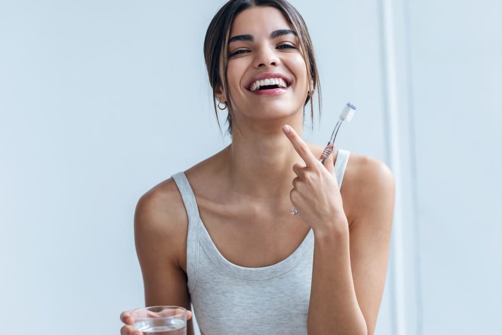 Woman Brushing Teeth With a Bright, Fresh Smile – Whitening A smiling woman holds a toothbrush and a glass of water, pointing to her teeth as she displays a clean, radiant smile achieved through daily care and whitening – Whitening.