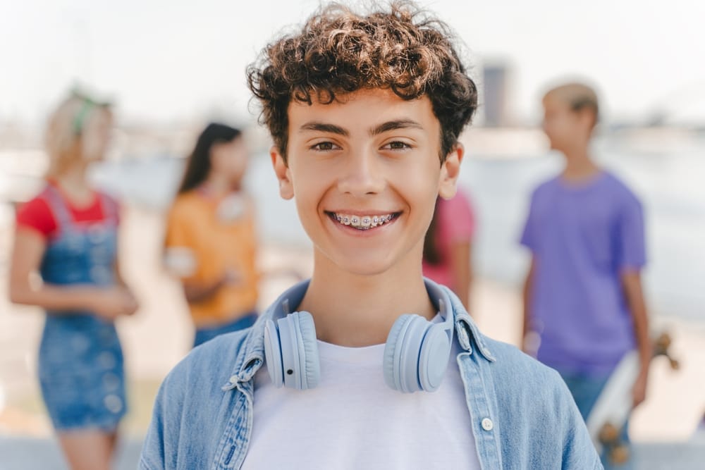 Smiling Teen Wearing Braces Outdoors – Braces in Denver A cheerful teen confidently smiles while wearing metal braces, showcasing the positive results of modern orthodontic treatment designed to create a straighter, healthier smile – Braces in Denver.