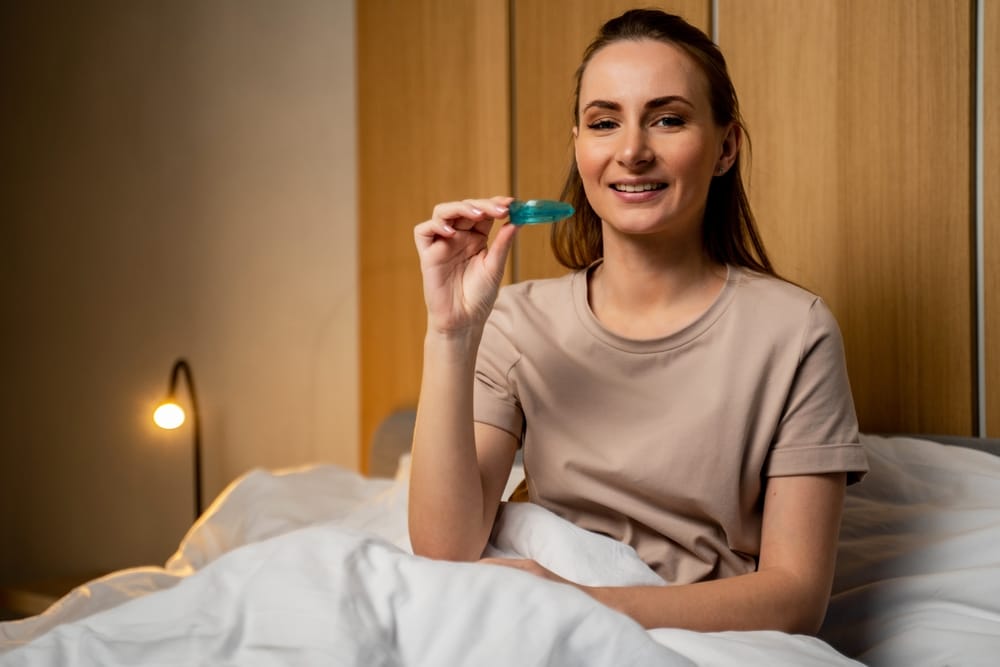 Woman Holding Night Guard Before Bed - Night Guards A woman sits comfortably in bed while holding a blue night guard, preparing to protect her teeth from nighttime grinding and clenching. - Night Guards