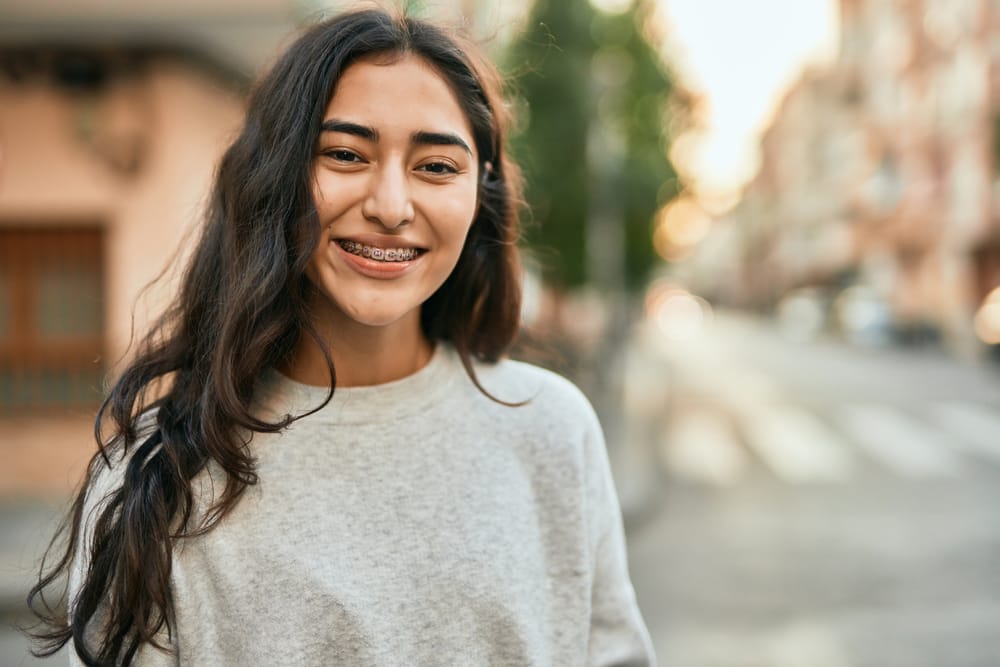 A young woman with long dark hair smiles while standing on a city street at sunset, wearing metal braces and a casual light gray sweatshirt – Denver Orthodontics & Dental Lounge