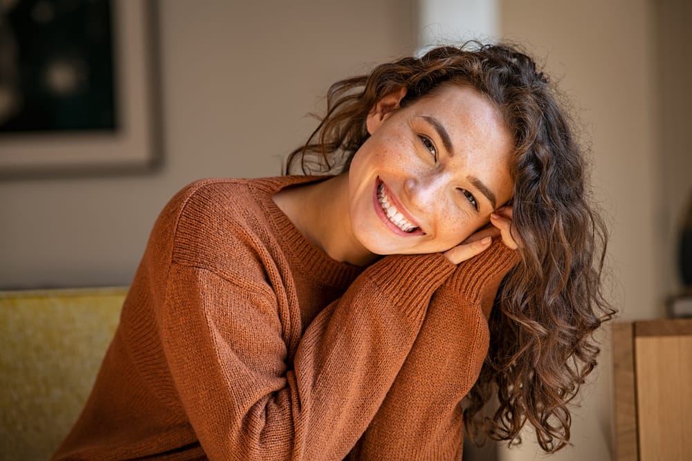 A cheerful young woman with curly hair rests her head on her hands, showing a warm and confident smile that reflects excellent orthodontic and dental care – Denver Orthodontics.